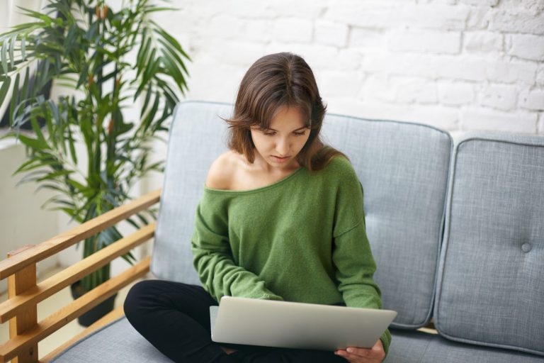 Eine Person in einem grünen Oberteil sitzt auf einem Sofa und schaut auf einen Laptop.